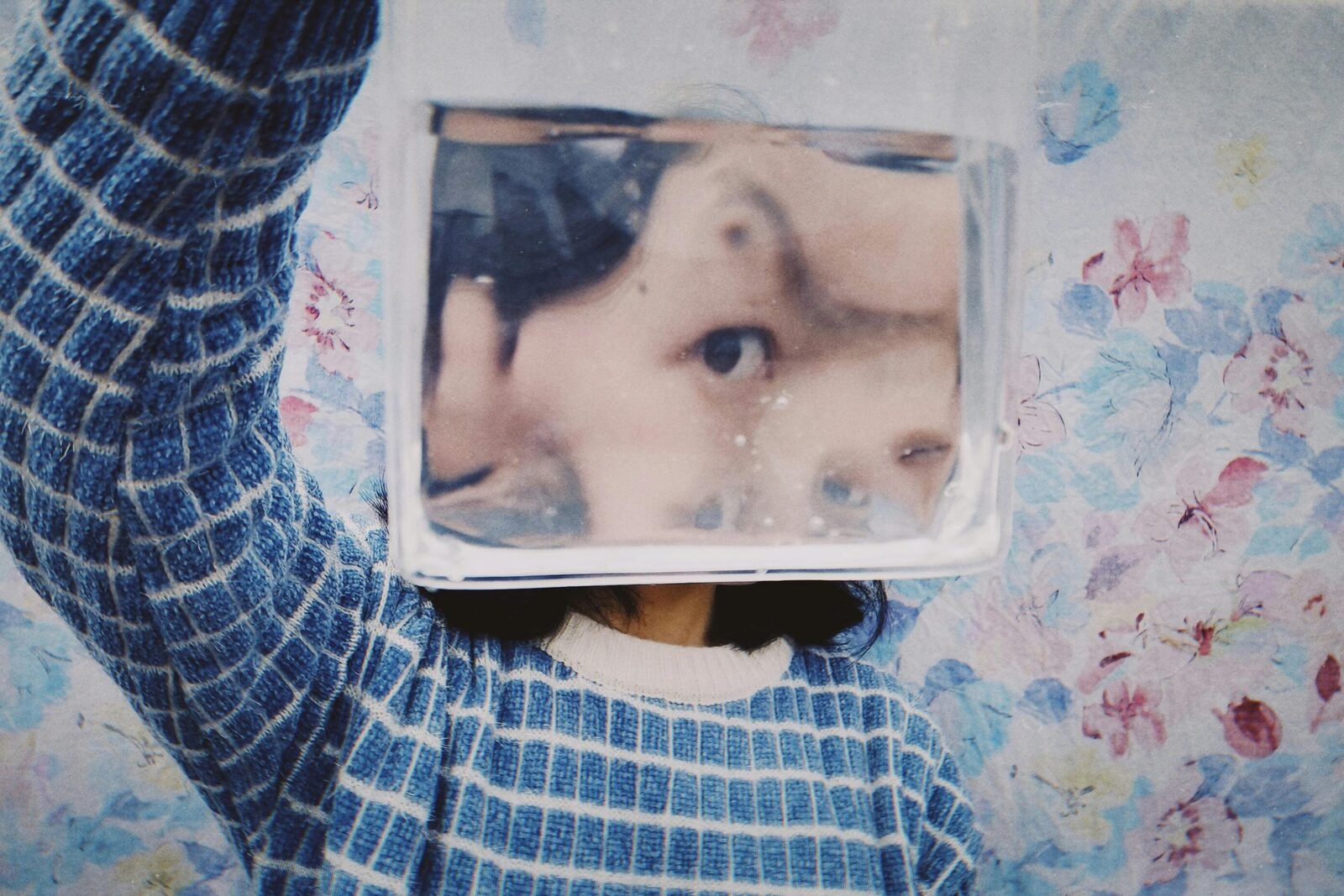 A person wearing a blue checkered sweater holds a clear container of water in front of their face, creating a surreal, distorted reflection of their features.